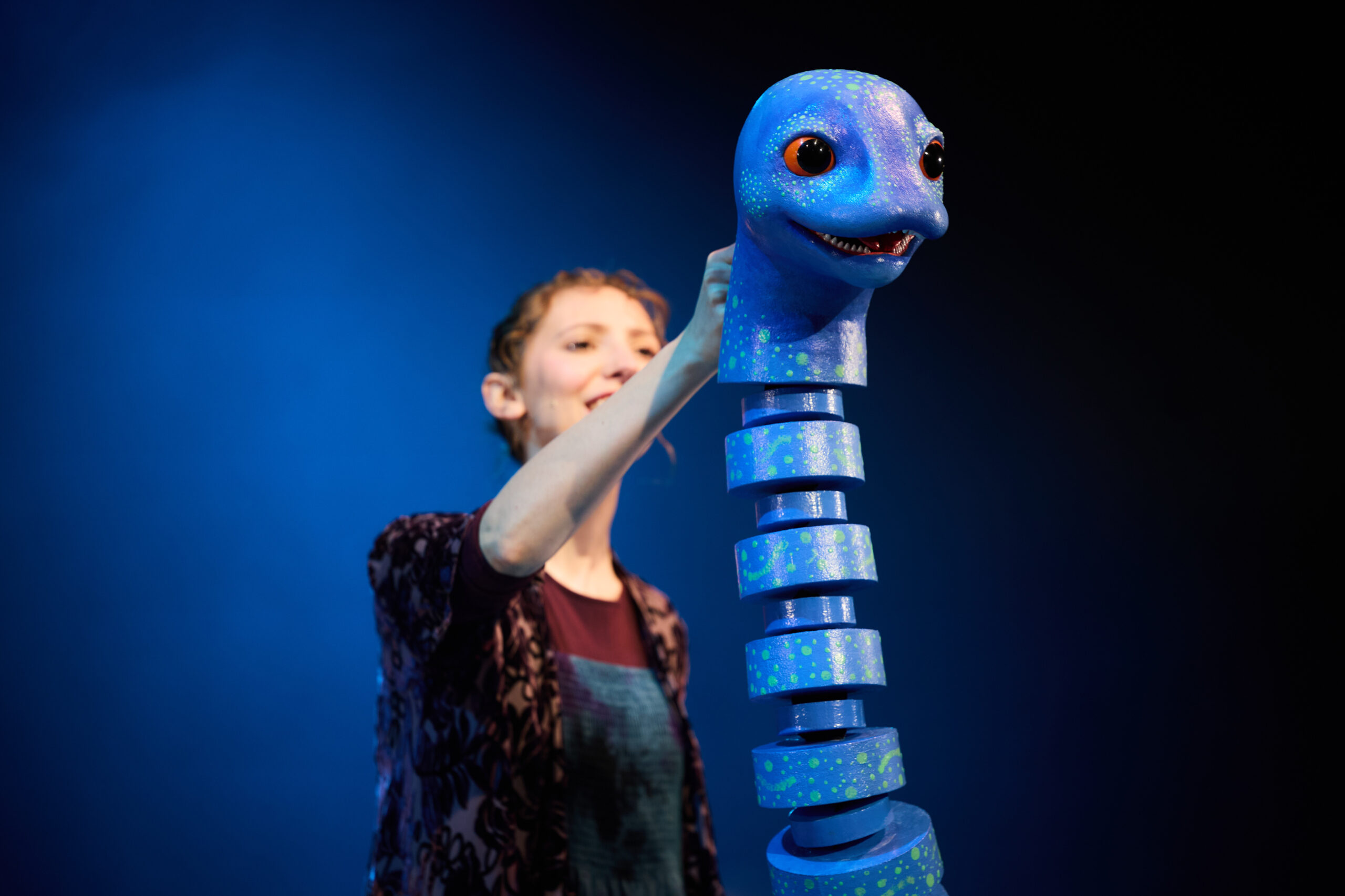 A person operates a segmented blue puppet resembling a loch ness monster against a blue background.