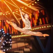 A ballet dancer in a gold costume performs a grand jeté on stage, with bright theater lights and a colorful background behind her.