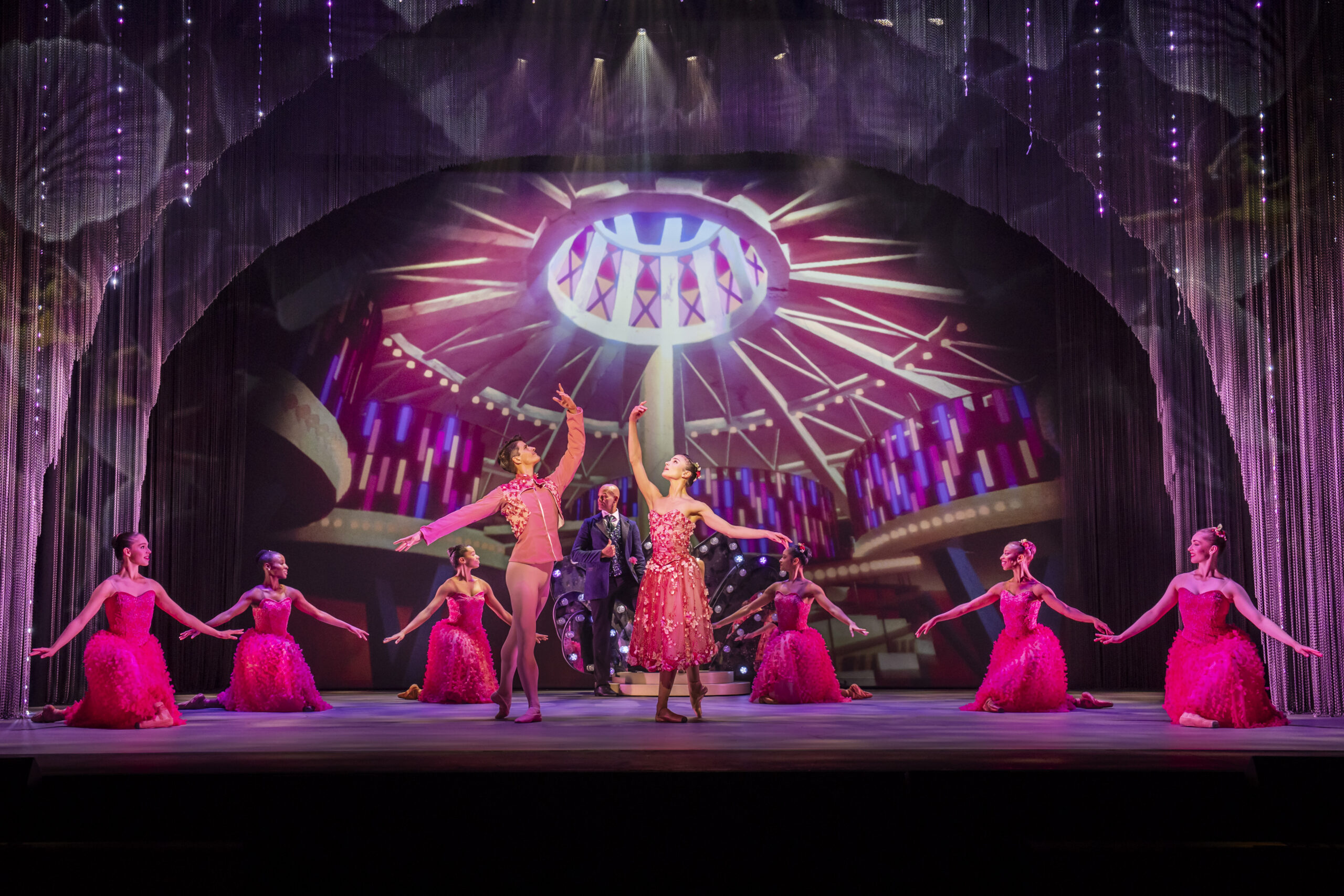 Dancers in bright pink costumes perform on stage with a dramatic circular light backdrop, while a man stands in the center under spotlights.