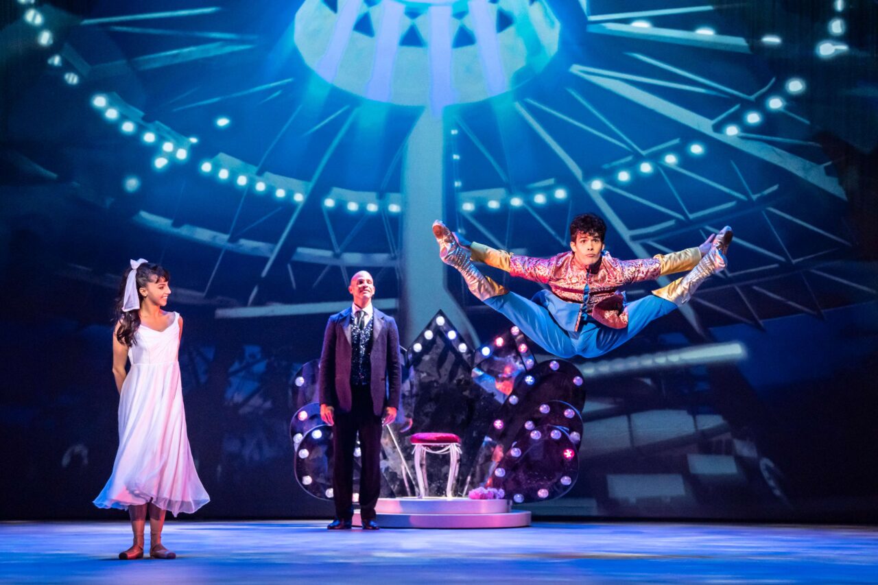 Three actors perform on stage; one leaps mid-air with arms and legs extended, while the other two stand in front of a throne-like chair under dramatic lighting.