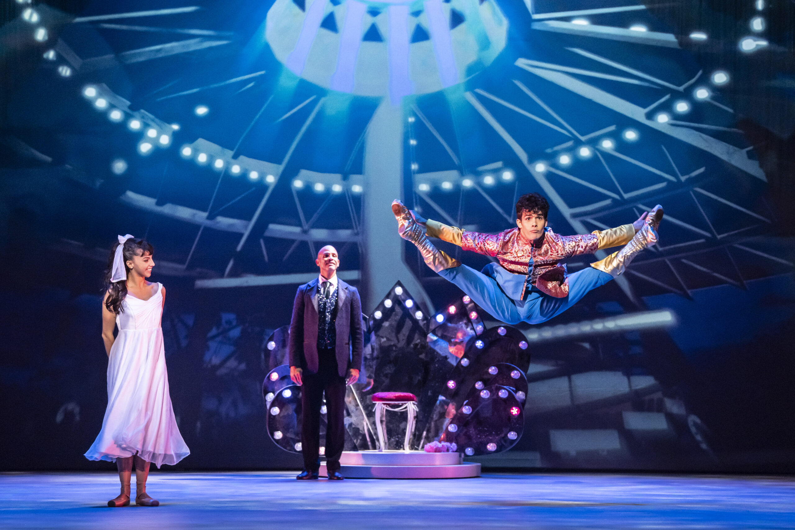 A performer in colorful attire jumps midair with legs extended, while a woman in a white dress and a man in a suit watch on a brightly lit stage.