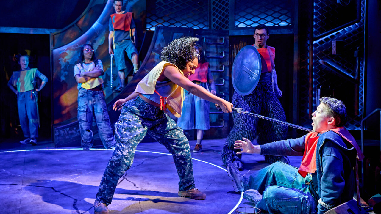 Actors on stage perform a dramatic scene where one person points a sword at another, while others watch in a brightly lit, industrial-themed set.