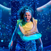 A woman wearing camouflage pants and a toilet seat around her neck shouts while holding two swords on a stage with blue lighting and bubbles in the background.