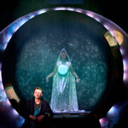 A person in contemporary clothing sits in front of a circular portal, behind which stands a figure in a glowing, hooded robe with a bright orb, surrounded by star-like lights.