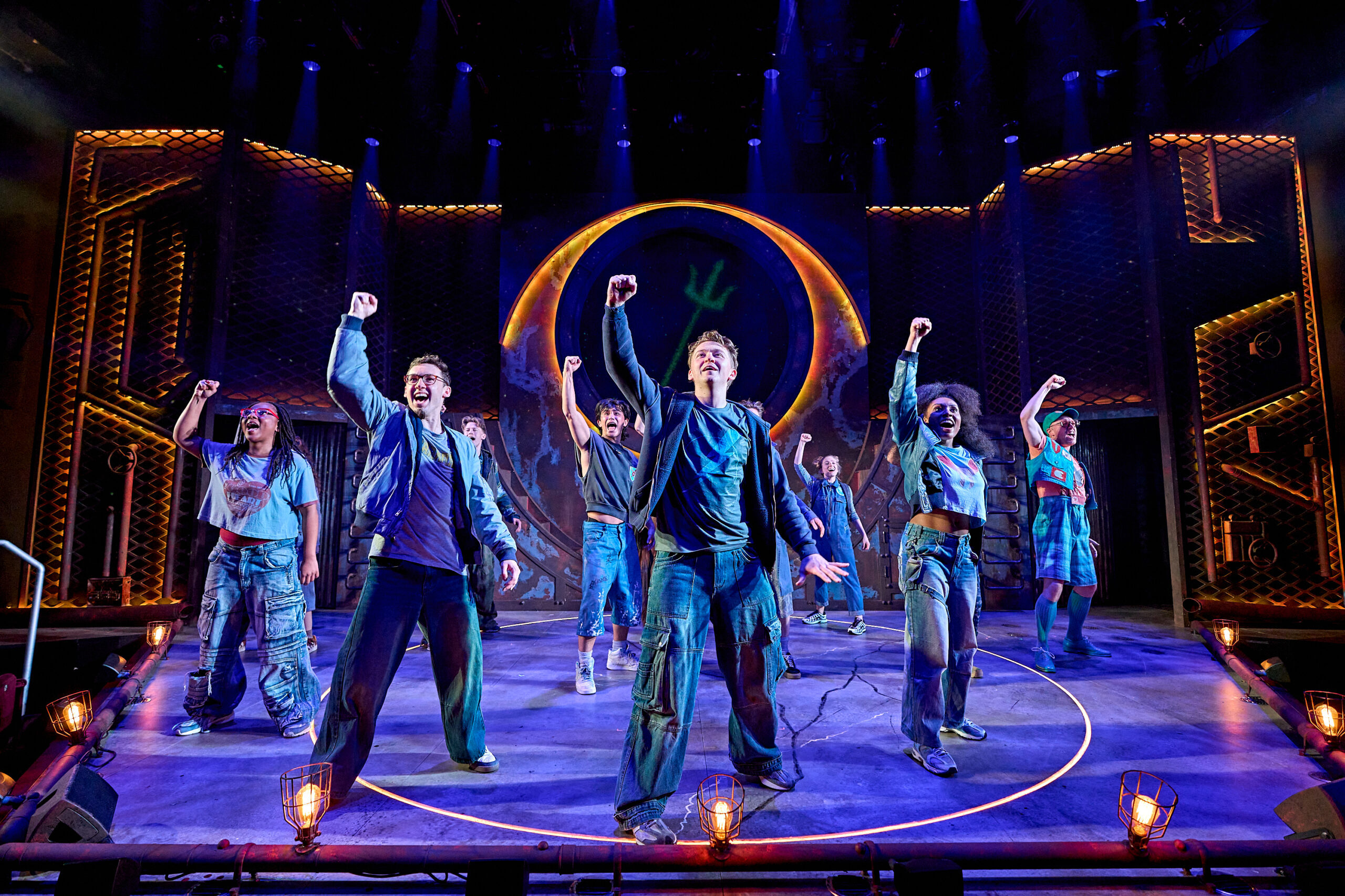 A group of actors on stage perform in front of a large circular set piece, each raising one fist in the air under dramatic blue and orange lighting.