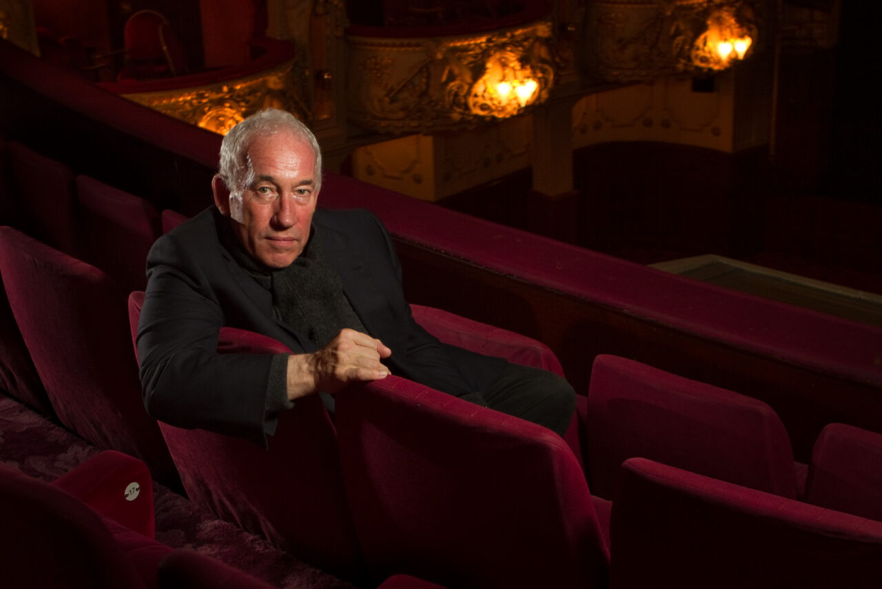 Simon Callow in a dark suit sits alone in a theatre's red velvet seats, with ornate balconies and warm lights visible in the background.