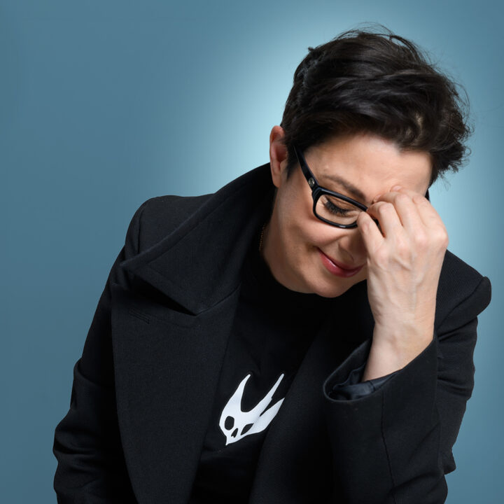 Person with short dark hair and glasses sits against a blue background, smiling with eyes closed and hand covering part of face.