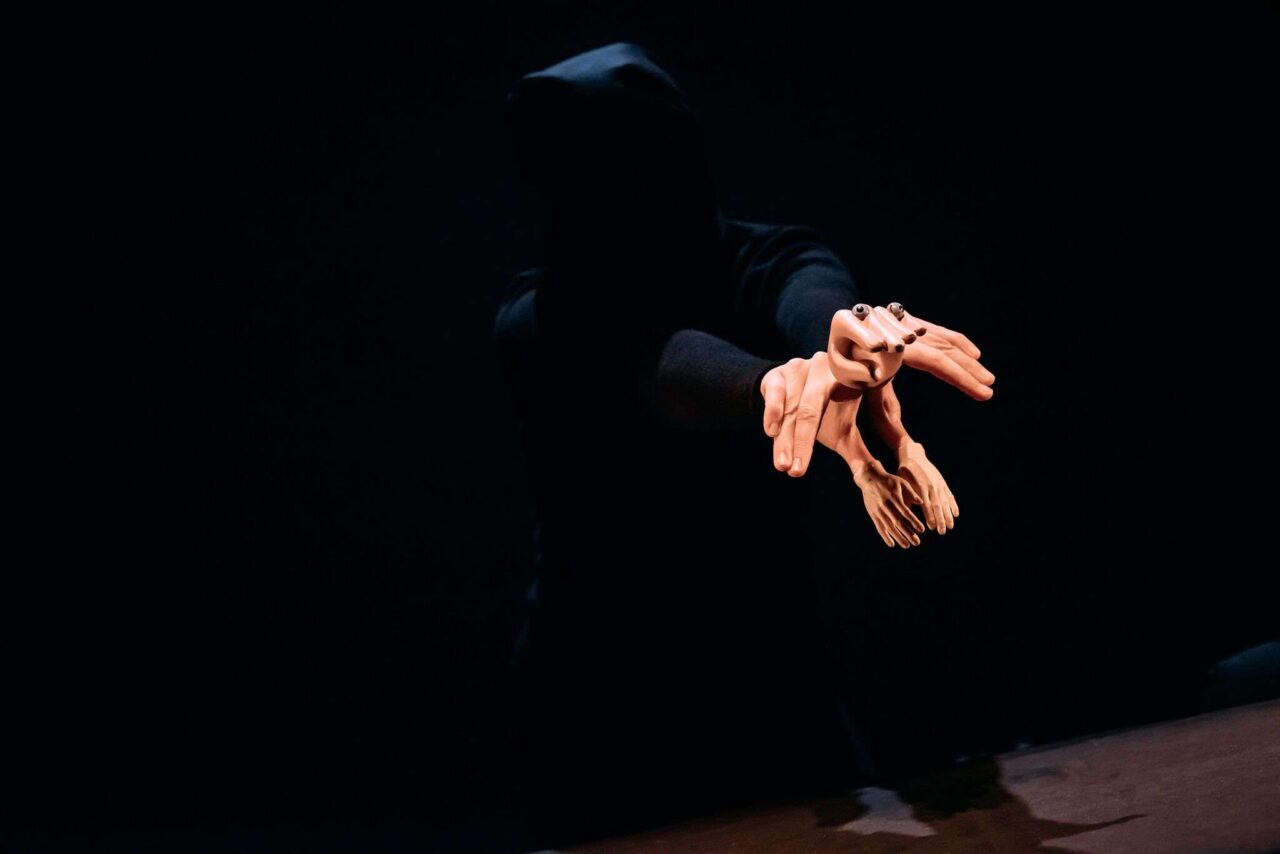 A person in a dark hoodie extends their arms forward, holding several small mannequin hands against a black background.