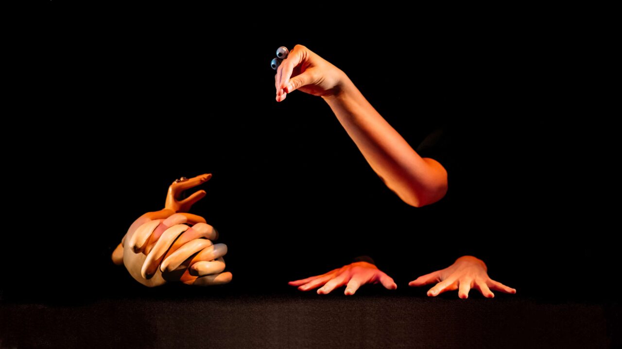 Four human hands are arranged against a black background; one holds a marble, two lie flat on a surface, and one set is clasped together.