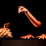 Four human hands are arranged against a black background; one holds a marble, two lie flat on a surface, and one set is clasped together.