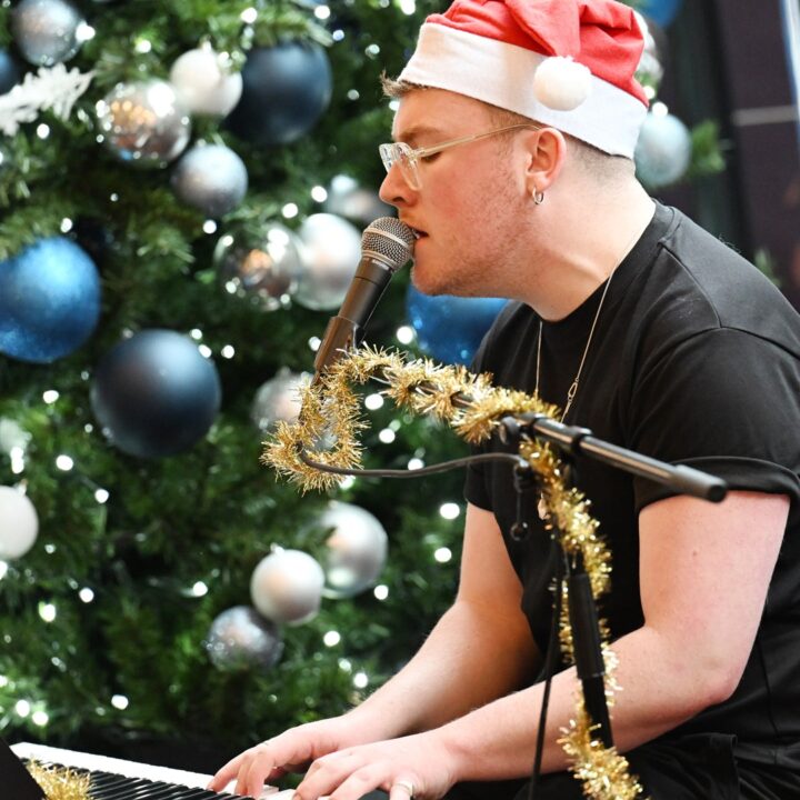Person in a Santa hat plays keyboard and sings, with a decorated Christmas tree and festive decorations in the background.