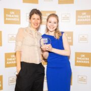 Two women pose for a photo at the UK Theatre Awards, one holding an award. They stand against a backdrop with the event's logo.