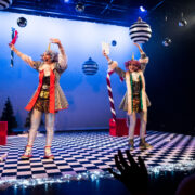 Two performers in colorful, clown-like costumes stand on a checkered stage decorated with oversized ornaments and candy canes, waving to an audience with hands visible in the foreground.