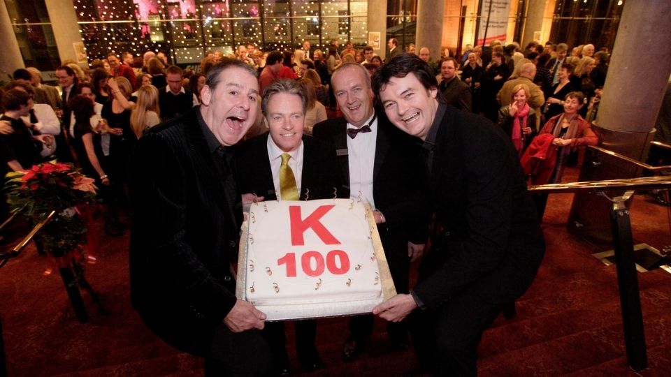 The panto cast hold a large cake with "K 100" written on it, standing on stairs in a crowded event venue decorated with lights.