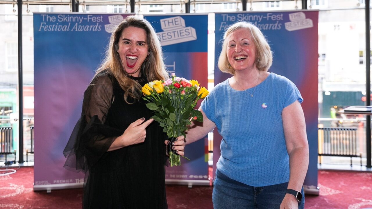 Two women smiling; one in black holding a bouquet of yellow and pink flowers, the other in blue. Event banners for "The Skinny-Fest Festival Awards" are visible in the background.