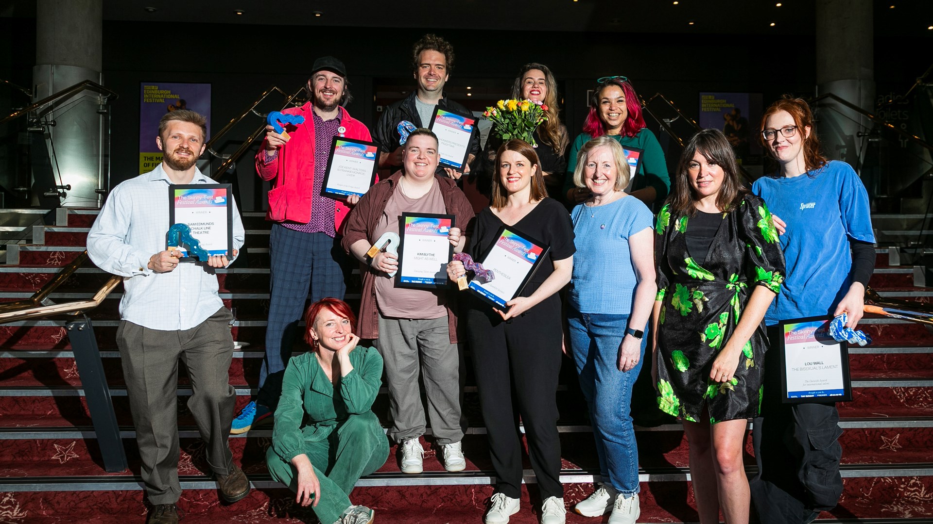 A group of eleven people pose on stairs indoors, some holding certificates and flowers, with a mix of casual and formal attire.