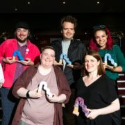 Seven people stand in two rows, each holding a colorful abstract-shaped award, posing and smiling for a group photo indoors under bright lighting.