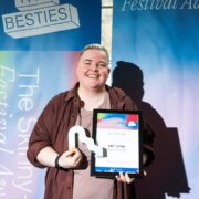 Person smiling and holding an award trophy and a framed certificate in front of a colorful "Festival Awards" backdrop.