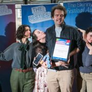 Six people pose together at The Skinny-Fest Festival Awards, with one holding a framed award and others displaying celebratory expressions in front of event banners.