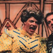Two actors, Allan Stewart and Andy Gray, in colorful costumes perform on stage, Allan is in Dame costume wearing a yellow polka-dot dress and wig, both have animated facial expressions against a painted backdrop.