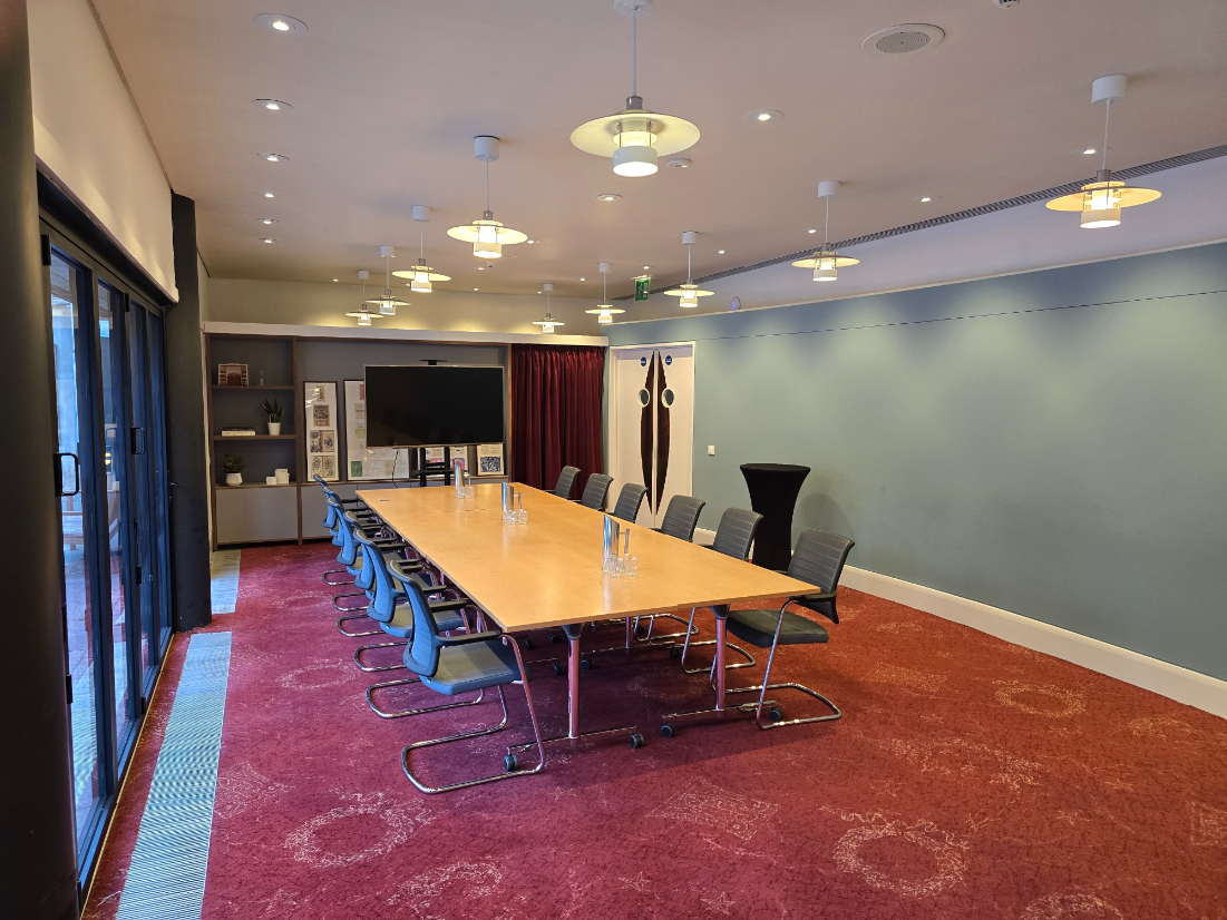 A modern conference room with a long wooden table, office chairs, water pitchers, a TV screen, and bookshelves, set on red carpet with overhead lighting.