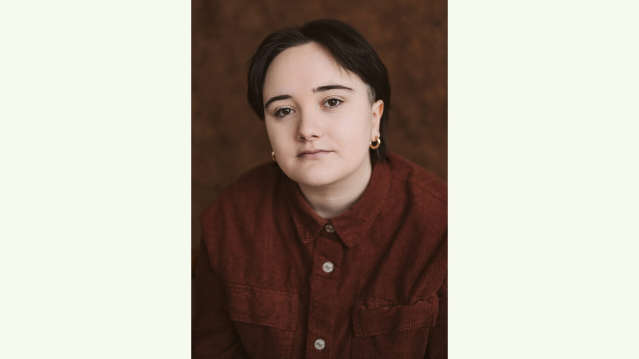 A person with short dark hair, wearing a red button-up shirt and small hoop earrings, looks directly at the camera against a brown background.