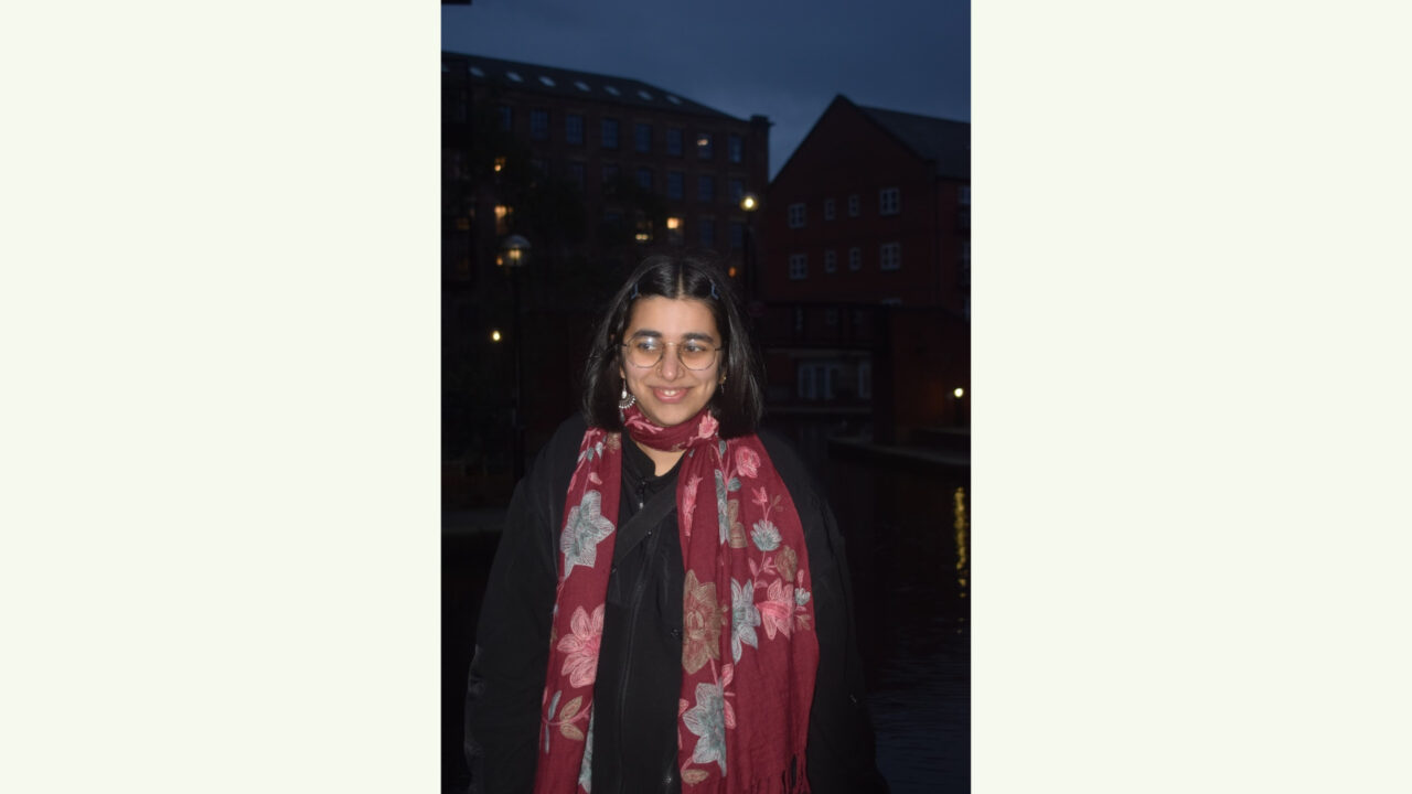 A person with glasses and a red floral scarf stands outside at night in front of buildings with lit windows.