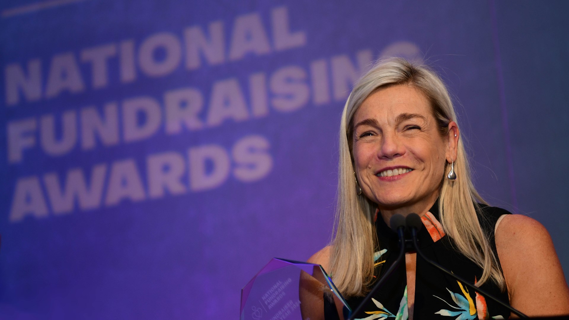 A woman smiles at a podium holding an award, with a "National Fundraising Awards" sign in the background.