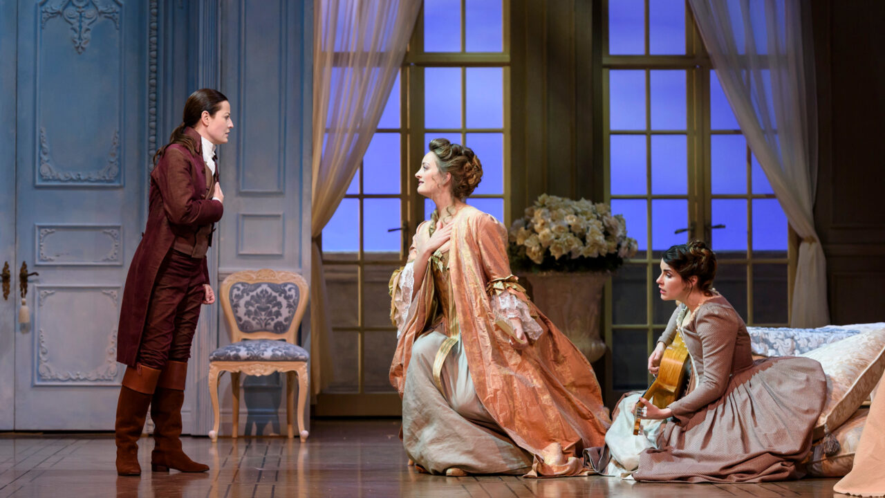 Three people in period costumes perform on a stage set evoking an elegant room with light blue walls and large windows—one stands, one kneels in a dress, and one sits on the floor holding a guitar, reminiscent of an Opera such as The Marriage of Figaro.