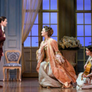 Three people in period costumes perform on a stage set evoking an elegant room with light blue walls and large windows—one stands, one kneels in a dress, and one sits on the floor holding a guitar, reminiscent of an Opera such as The Marriage of Figaro.