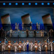 A large cast performs a musical number on a two-level stage set resembling a ship, with lifebuoys, sailor costumes, and smokestacks in the background.