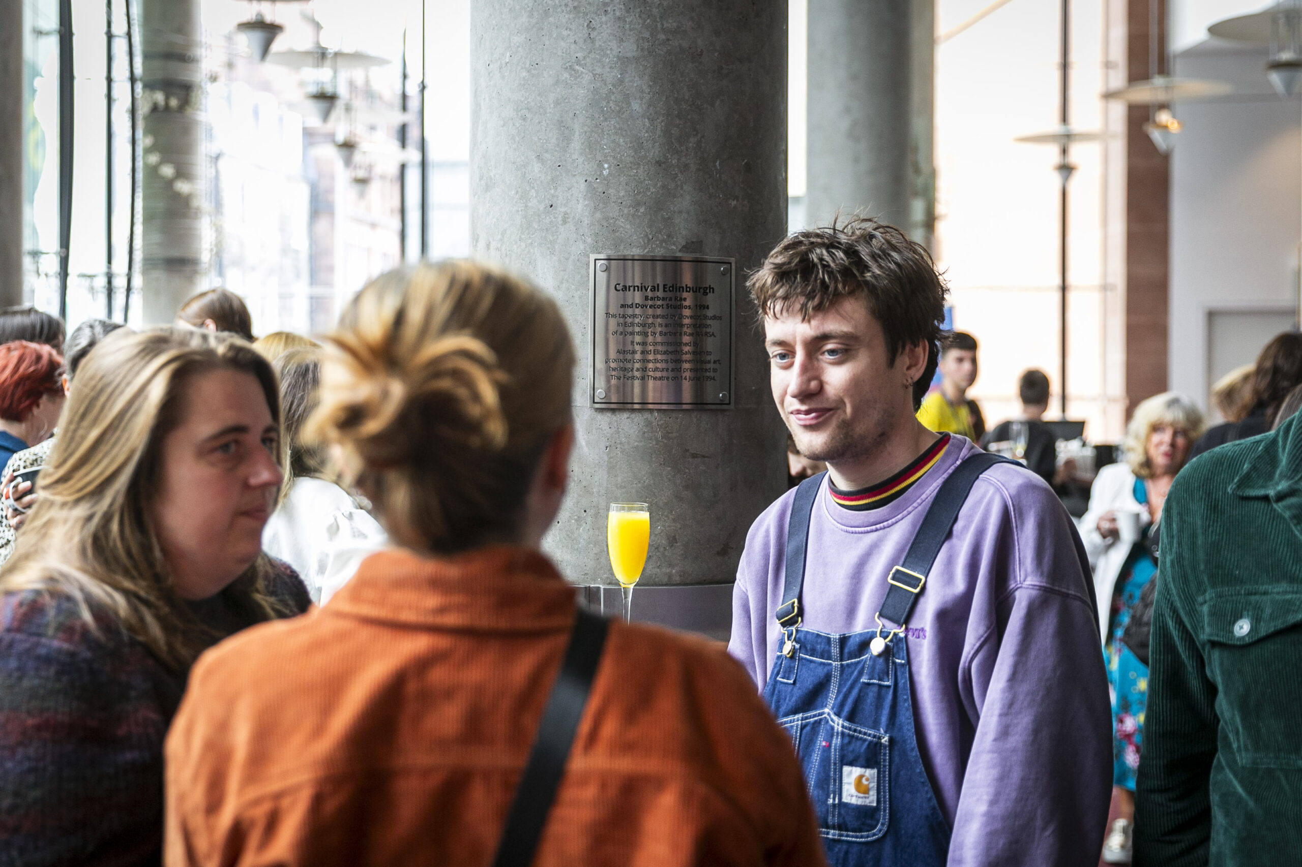 A group of people stand indoors having a conversation; one person in the foreground wears a purple sweatshirt and overalls, with a drink on a table nearby.