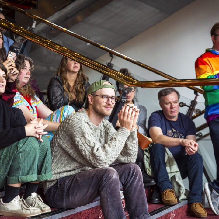 A group of people sit and stand on stairs, attentively watching something off-camera. One person stands wearing a rainbow-colored jacket.