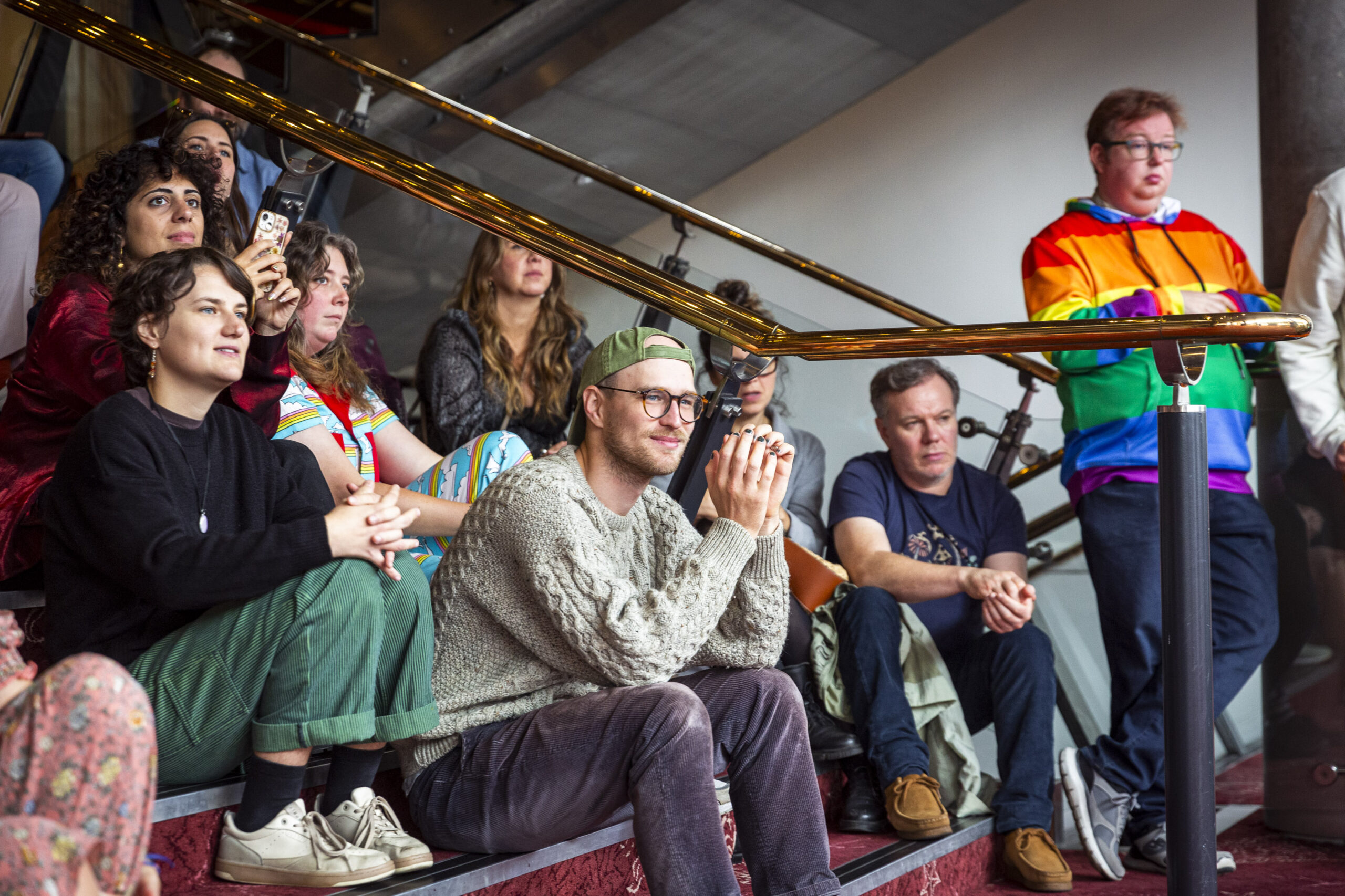 A group of people sit and stand on stairs, attentively watching something off-camera. One person stands wearing a rainbow-colored jacket.