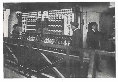 Black and white photo. Three people, including Bethia on the far left, stand in front of a large early 20th-century electrical control panel.