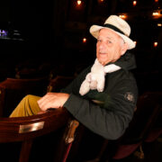 An older man wearing a hat and scarf sits in an empty theatre, looking back over his shoulder toward the camera.