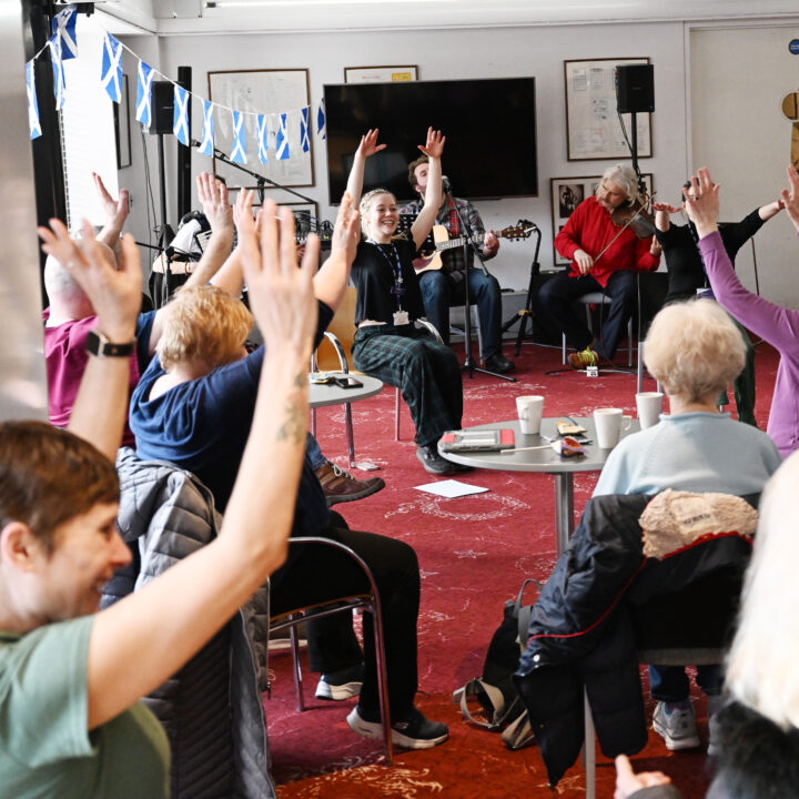 A group of older adults sit in a semicircle raising their arms during an indoor group activity; a few musicians are playing instruments at the front.
