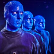 Three people with blue-painted faces and bald heads wearing black outfits stand in a row against a dark background.