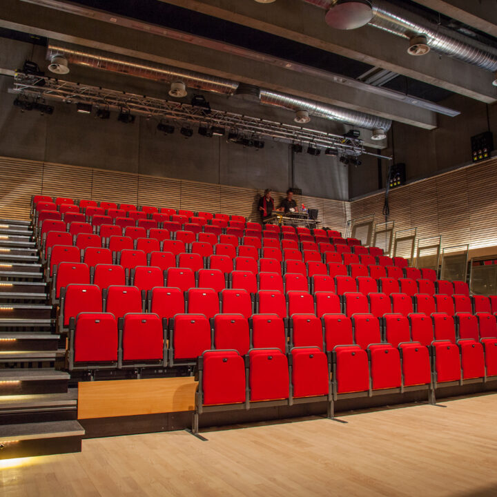 A modern auditorium with tiered rows of red seats, wooden floor, and exposed ceiling pipes, viewed from the stage area.