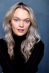 A woman with long wavy blonde hair and blue eyes is wearing a black turtleneck top, posing against a plain blue-gray background.