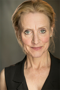 A middle-aged woman with light hair pulled back, wearing a black sleeveless top, looks directly at the camera with a neutral expression.