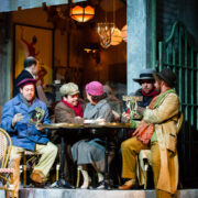 Five people in vintage attire sit at an outdoor café table, reading papers, with a waiter and another person visible inside the warmly lit restaurant.