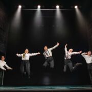 Five people in dress shirts and suspenders are mid-jump, performing a synchronized dance routine on a stage under bright stage lights.
