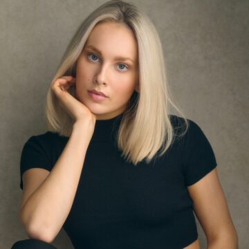 A woman with straight blonde hair, wearing a black short-sleeve crop top and black pants, sits with her hand resting on her chin against a neutral background.