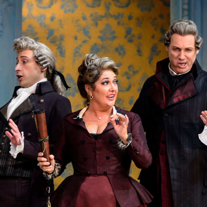Three people in 18th-century costumes perform on stage, with expressive gestures, in front of a blue and yellow patterned backdrop.