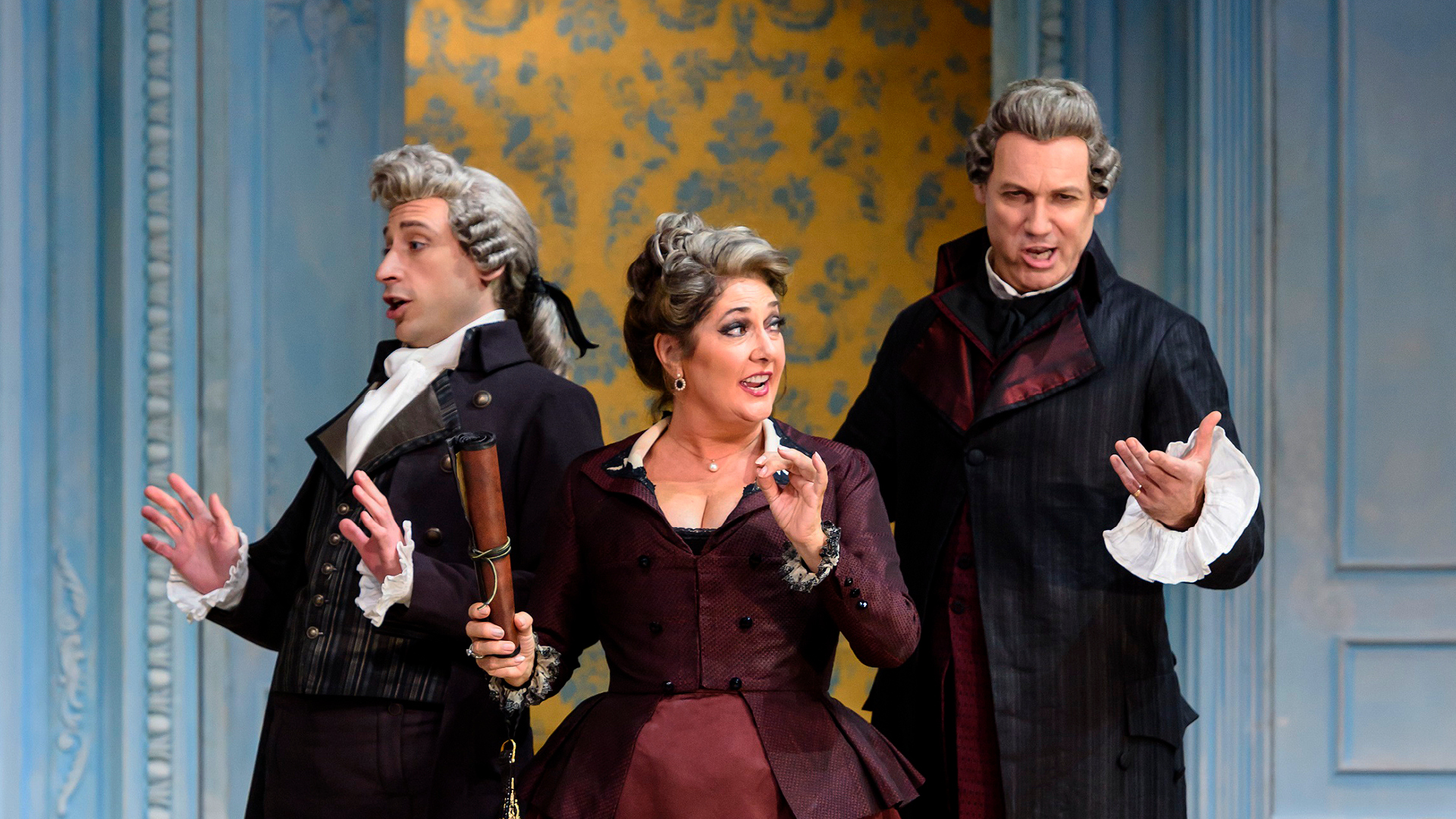 Three people in 18th-century costumes perform on stage, with expressive gestures, in front of a blue and yellow patterned backdrop.