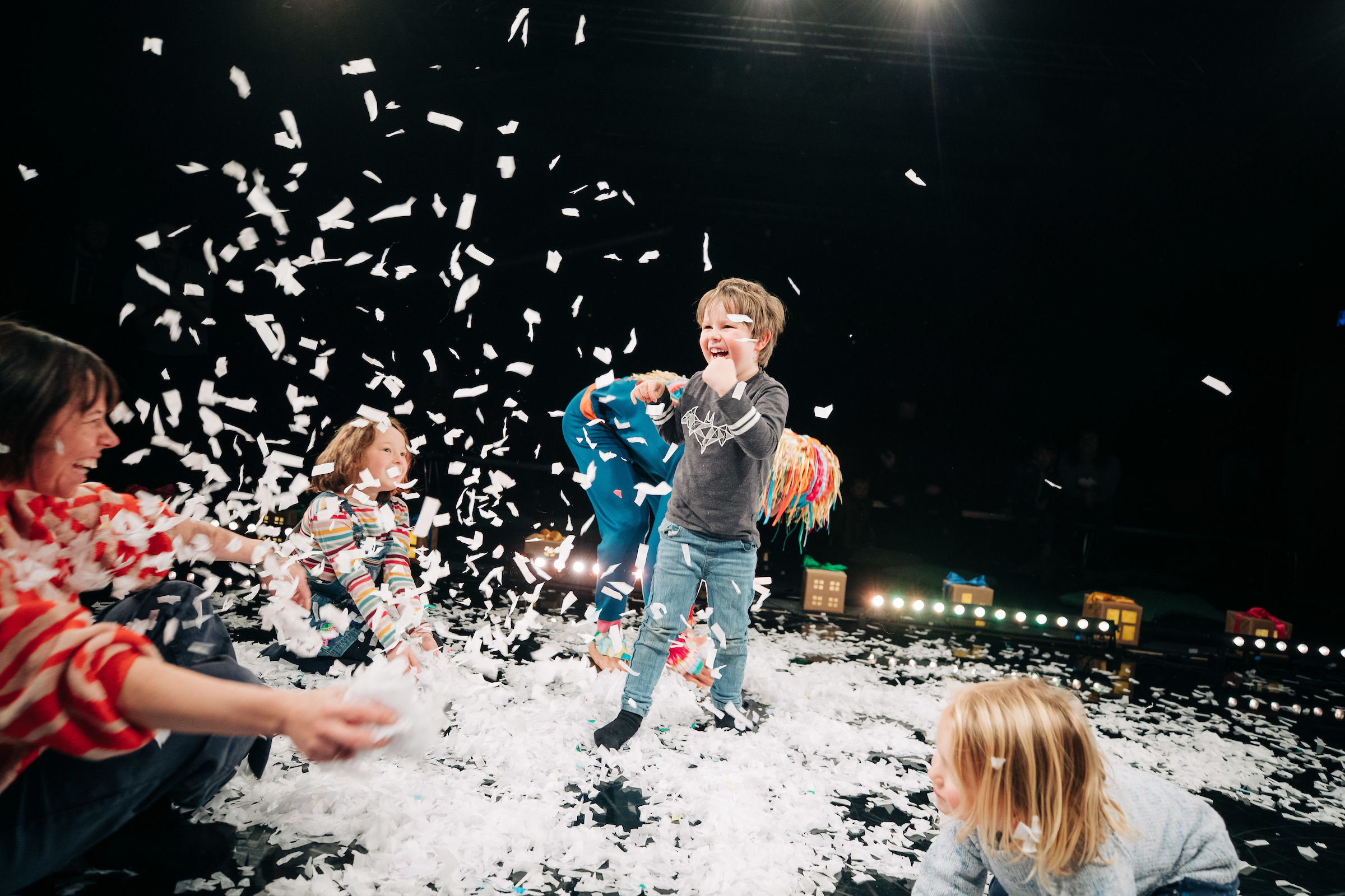 Children play and laugh as white paper confetti falls around them on a stage, with adults joining in the activity.