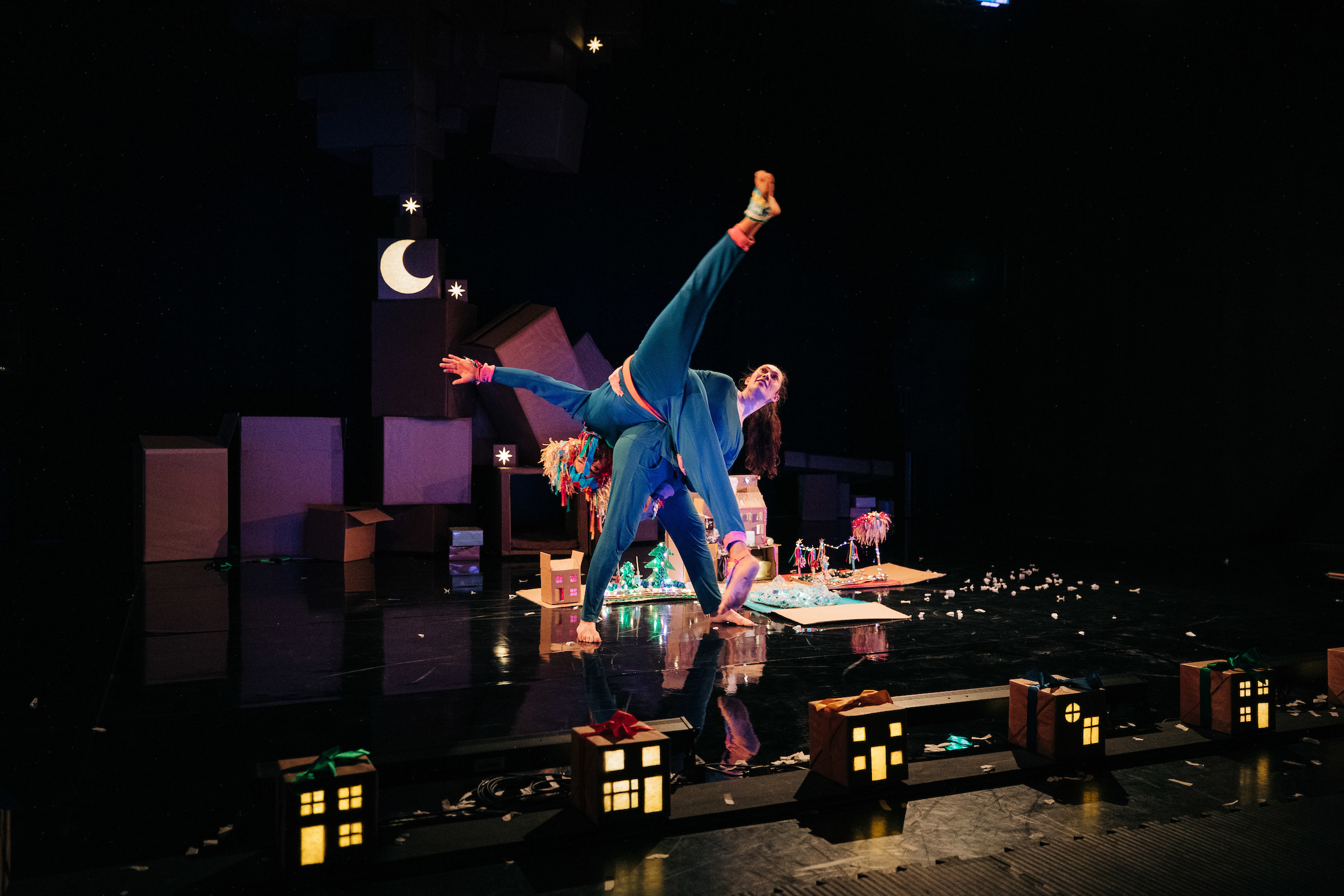 Two performers in colorful costumes do an acrobatic pose on stage, surrounded by cardboard props and small illuminated house models, under dim lighting with a crescent moon backdrop.