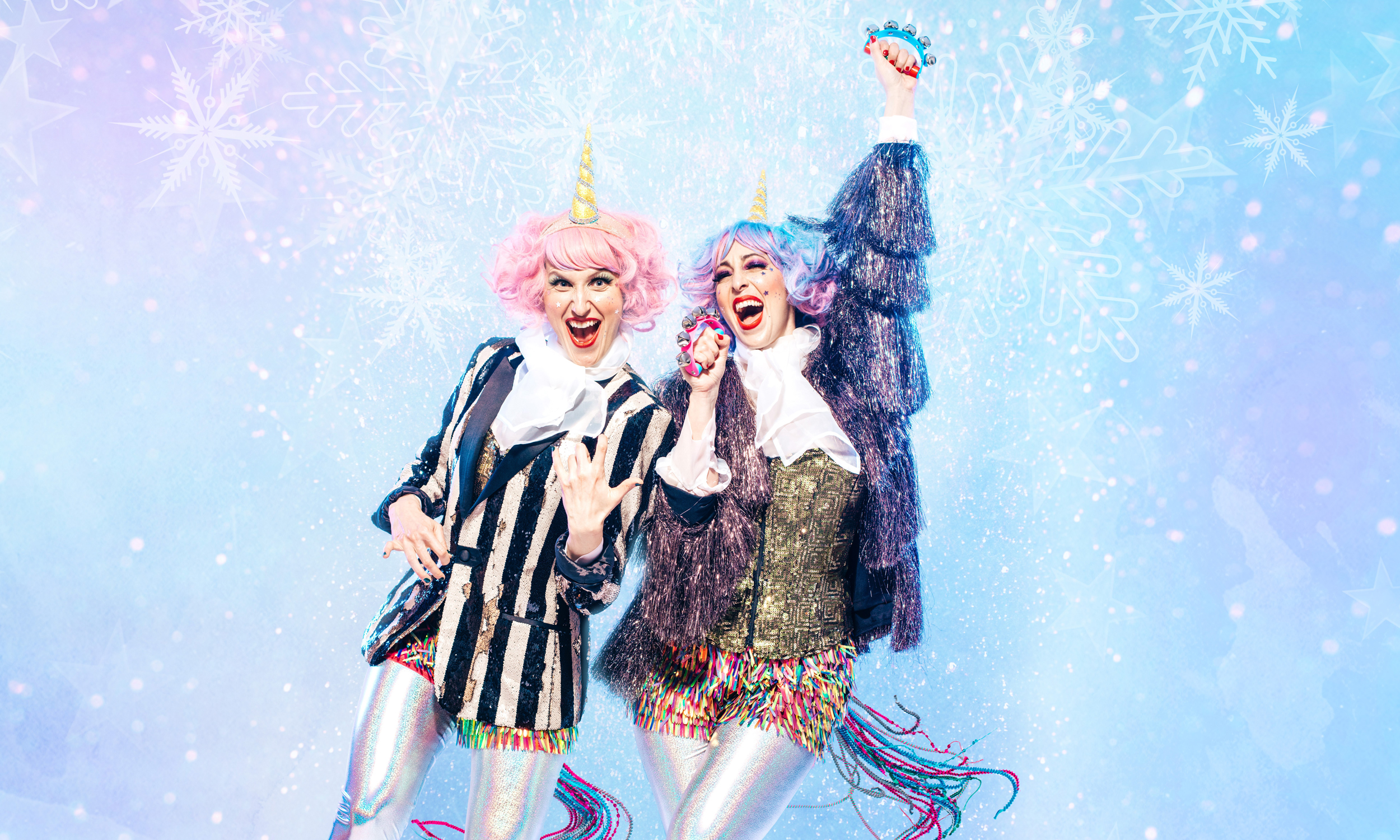 Two people dressed in colorful unicorn-themed costumes with pink and purple wigs, striped jackets, and sparkly shorts stand smiling and posing against a blue, snowy, festive background.