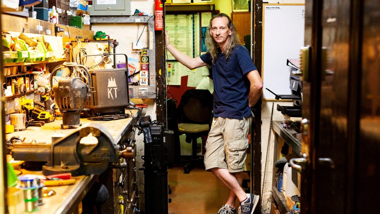 A man with long hair, wearing a navy shirt and khaki shorts, stands in a cluttered workshop with tools and equipment on shelves and workbenches.